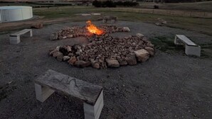 Ein runder Feuerkorb aus Stein mit Feuer, umgeben von Bänken und einer Wiese.