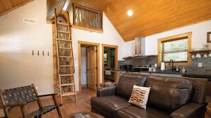 Un interior acogedor con techo de madera, un sofá de cuero marrón, una escalera de madera y una zona de cocina con salpicadero de ladrillo.