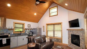 Un espacio de sala acogedor con chimenea, un revestimiento de piedra alrededor de la chimenea, un sofá de cuero y una cocina con refrigerador de acero inoxidable.