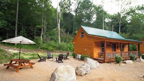 Una cabaña de madera con porche, rodeada de un área de grava con mesas de picnic y sillas Adirondack.