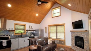Un espacio de sala acogedor con chimenea, un revestimiento de piedra alrededor de la chimenea, un sofá de cuero y una cocina con refrigerador de acero inoxidable.