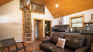 Een knus interieur met een houten plafond, een bruine leren bank, een houten ladder en een keuken met een tegelachtergrond.
