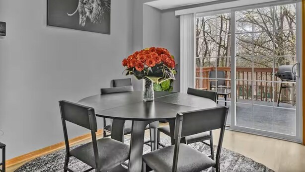 Un comedor con una mesa redonda, seis sillas, un jarrón con flores rojas, una obra de arte negra en la pared y un ventanal con vistas a árboles.