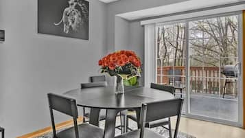 Een eetruimte met een ronde tafel, zes stoelen, een vaas met rode bloemen, een zwart kunstwerk aan de muur en een groot raam met uitzicht op bomen.