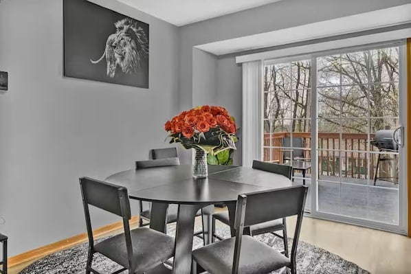 A dining area with a round table, six chairs, a vase with red flowers, a black artwork on the wall, and a large window with a view of trees. - Cozy stylish home near Akron ~Canton/ sleeps 8 (Akron)