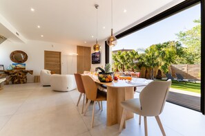 A modern dining area with a wooden table, chairs, and a view of a garden with palm trees. - Villa Amphitrite • Share, beach & happiness (Agde)