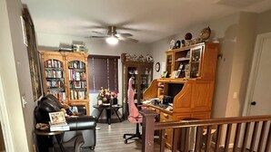 Un interior de vivienda con una escalera de madera, un ventilador de techo, una estantería, un escritorio, una silla y otra estantería repleta de libros.