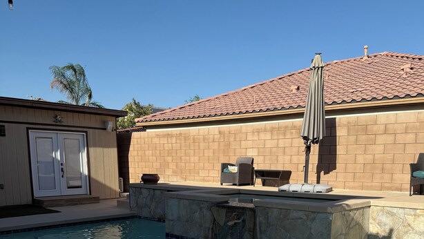 Una piscina con borde de piedra, un patio cubierto con mobiliario exterior y un edificio con tejado de azulejos.