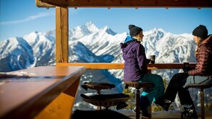 Zwei Personen sitzen an einem Holztisch mit Blick auf schneebedeckte Berge.