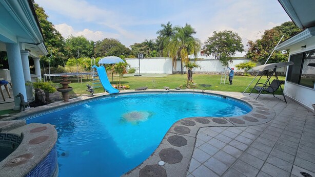 A swimming pool with a blue slide, a stone deck, and a grassy yard with palm trees. - Villa Cocoyoc for 23 Pax | Pool, Jacuzzi and Basketball Court (Fraccionamiento Lomas de Cocoyoc)