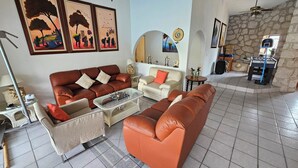 A living room with a brown sofa, a white sofa, a glass coffee table, and a red armchair. There are framed pictures on the wall and a stone wall in the background. - Villa Cocoyoc for 23 Pax | Pool, Jacuzzi and Basketball Court (Fraccionamiento Lomas de Cocoyoc)