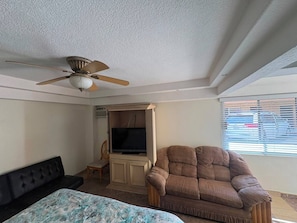 A room with a ceiling fan, a television, a sofa, a chair, and a window with blinds.