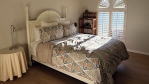 Quarto com cama, luminária de cabeceira, mesa redonda, ventilador de teto e janela com persianas.