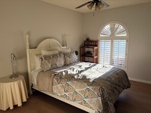 A bedroom with a bed, a bedside lamp, a round table, a ceiling fan, and a window with blinds. - Spacious 3-bedroom Home for Rent in 55+ Active Adult Community (Sun Lakes)