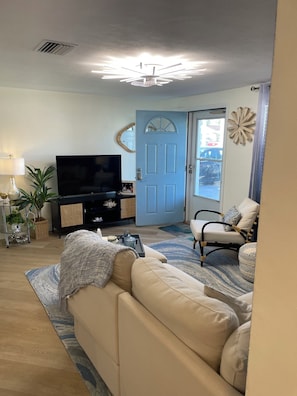 A living room with a beige sofa, a flat-screen TV, a decorative mirror, and a door with a unique design. - Cozy chic 2-bedroom house in Port Richey with screened in pool (Port Richey)