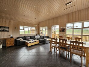 A room with a wooden ceiling, a sofa, a coffee table, a dining table with chairs, and large windows with a view.