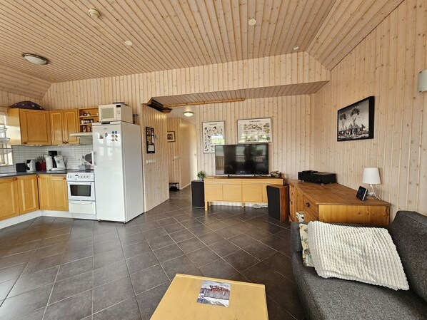 A modern kitchen with wooden cabinets, a white refrigerator, and a stove. There is a dining area with a wooden table and chairs, a TV, and framed pictures on the wall.