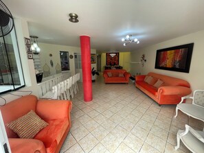 Una sala de estar con sofás naranja, un pilar rojo, una zona de comedor con sillas blancas y un ventilador de techo.