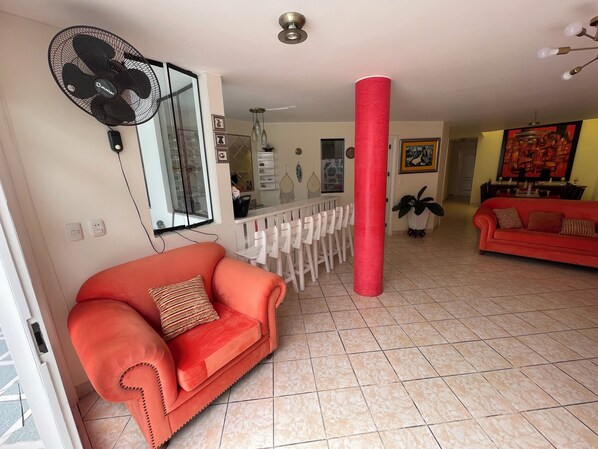 Una sala de estar con un sofá rojo, un ventilador, un comedor con sillas, un pilar rojo y un televisor al fondo.