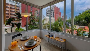 Een kamer met een ontbijt aan tafel gedekt, een bankje met een krant, en grote ramen met uitzicht op gebouwen en groen.