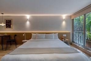 A modern hotel room with a large bed, a desk, a chair, a small table, a lamp, and a large window with a view of greenery.