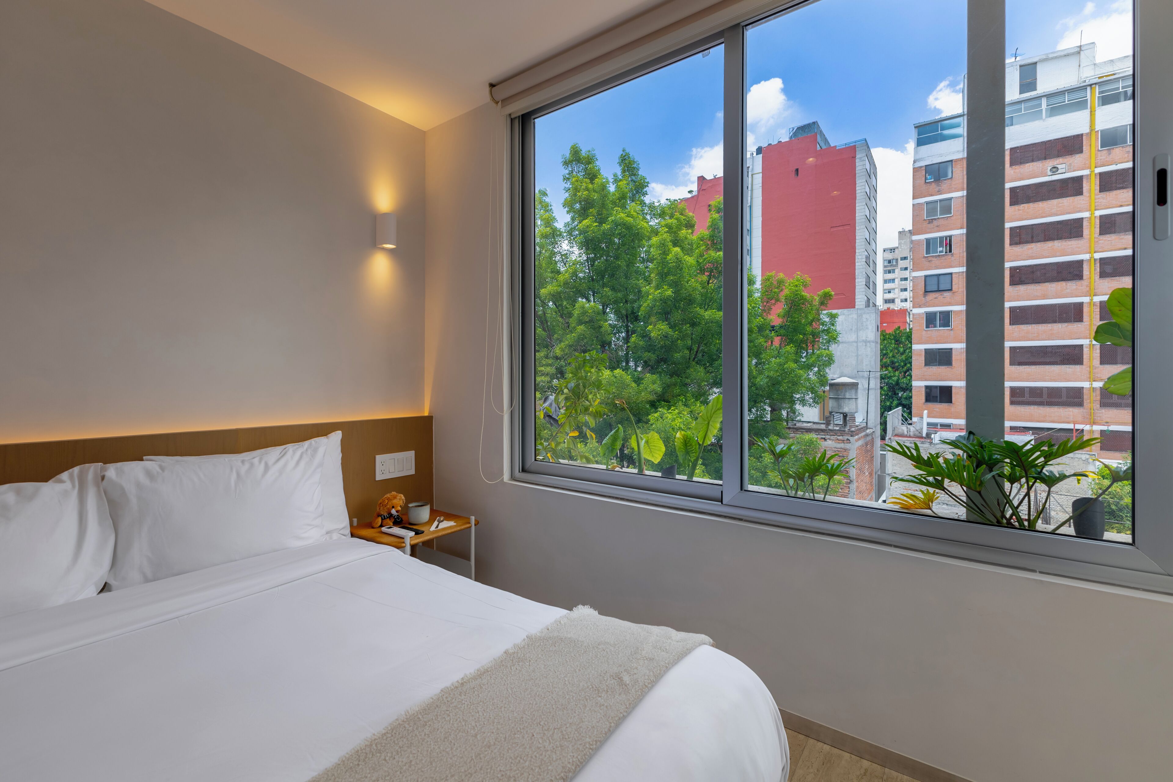 Ein Hotelzimmer mit großem Fenster, einem Bett, einem Nachttisch und einer Wandleuchte.