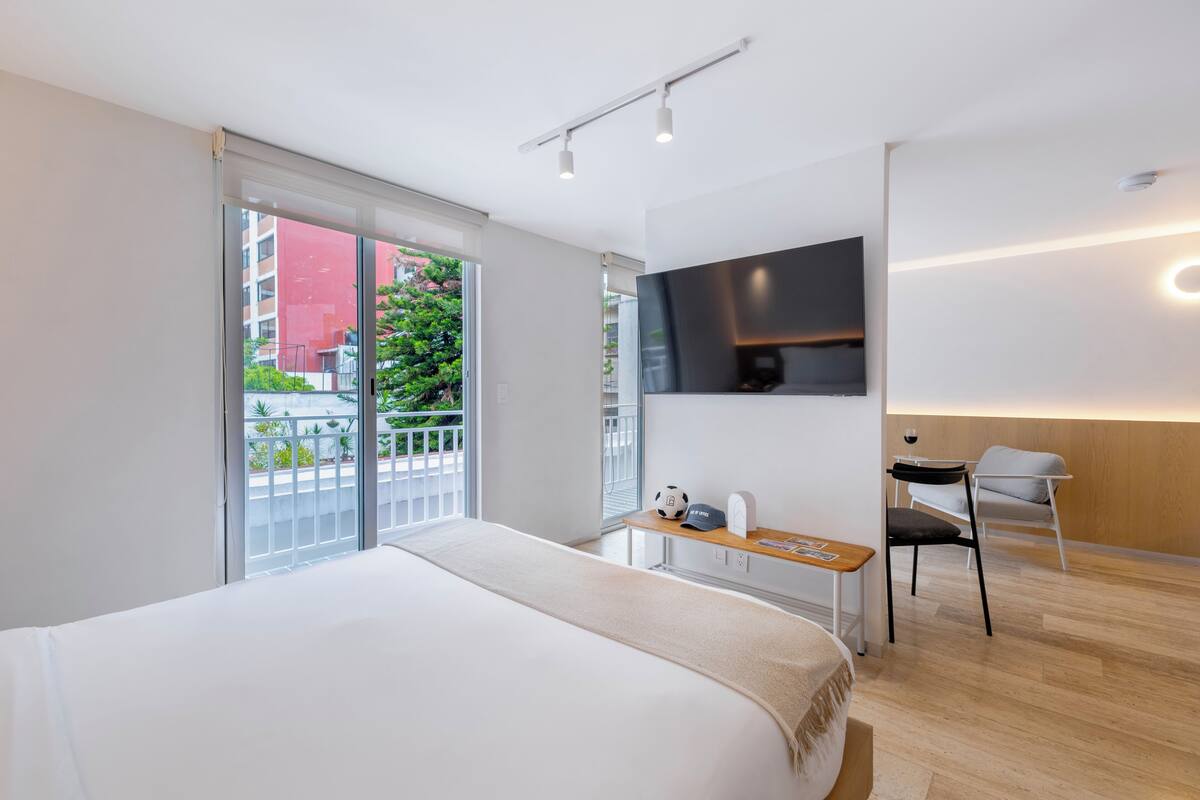 Habitación de hotel moderna con una cama grande, un escritorio con silla, un televisor en la pared y un balcón con vistas a edificios y vegetación.