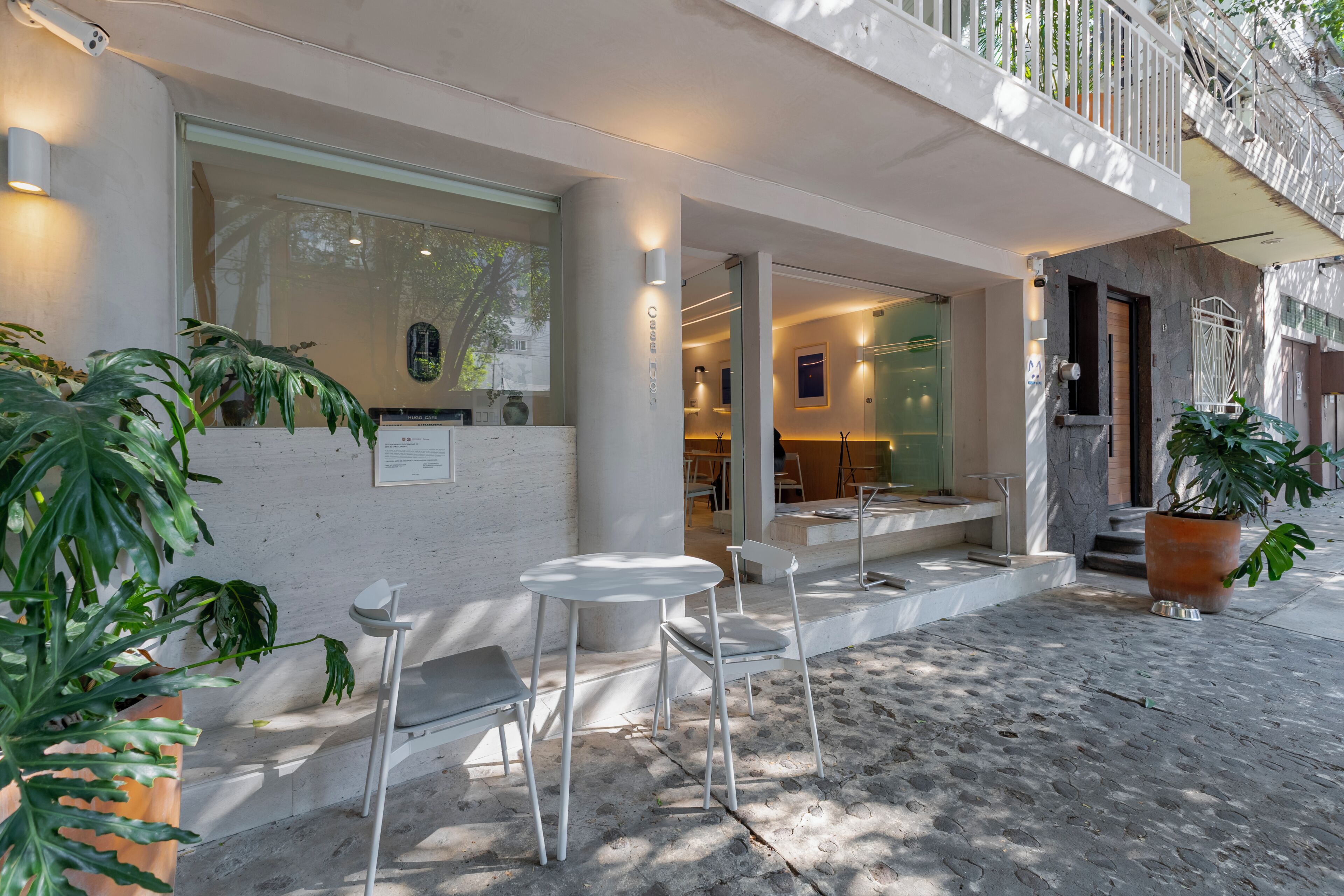 Un espace extérieur moderne aménagé avec des chaises et des tables blanches, des plantes en pot et une vue sur l’intérieur à travers une porte vitrée.
