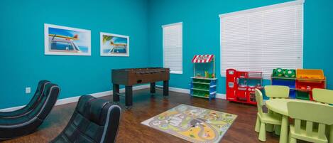 Une pièce aux murs de couleur turquoise, avec un baby-foot, une table de billard et un espace de jeux équipé de jouets et d’une petite structure rayée rouge et blanche.