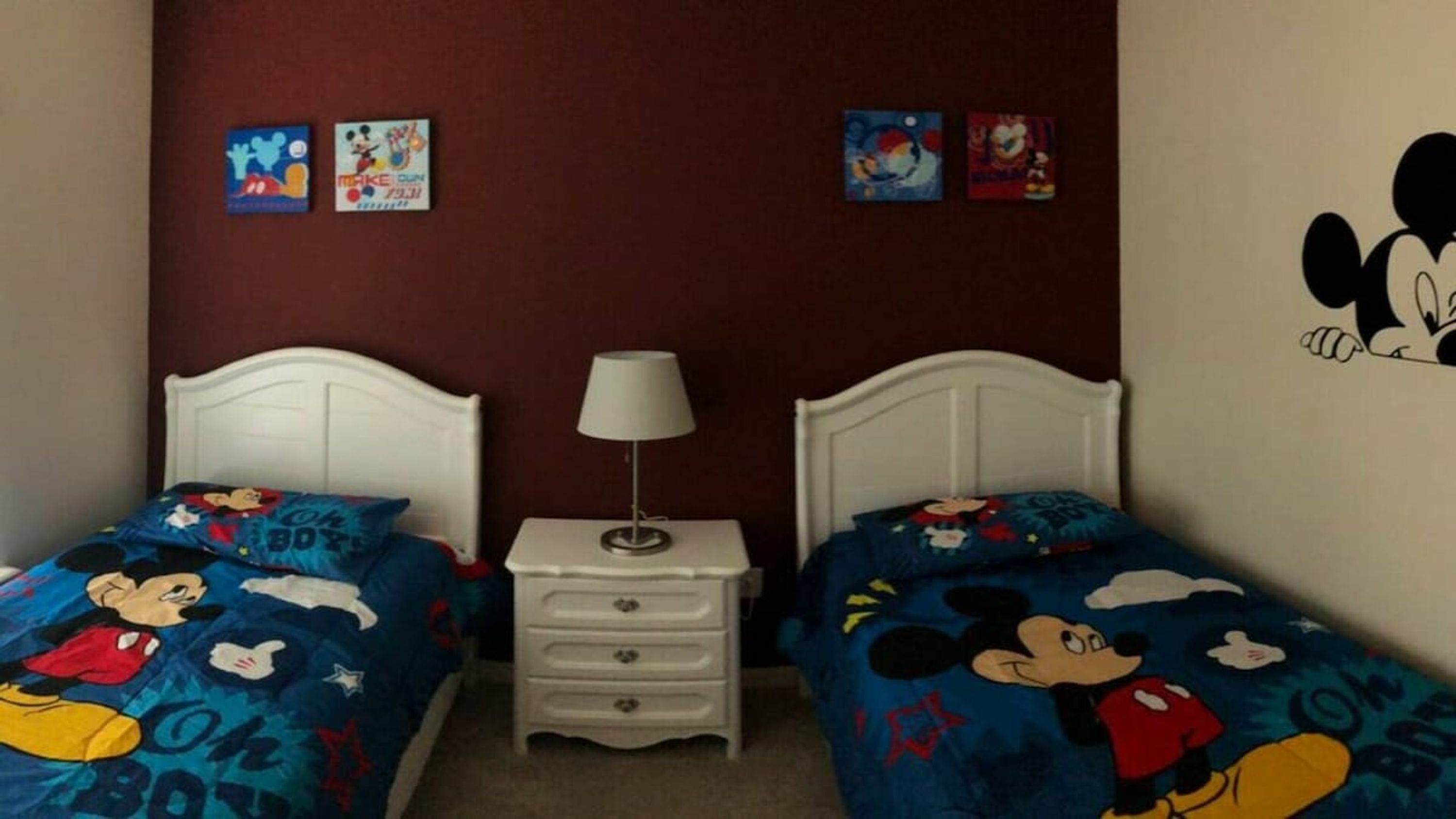 A bedroom with two beds, each with Mickey Mouse bedding, a nightstand with a lamp, and wall art.