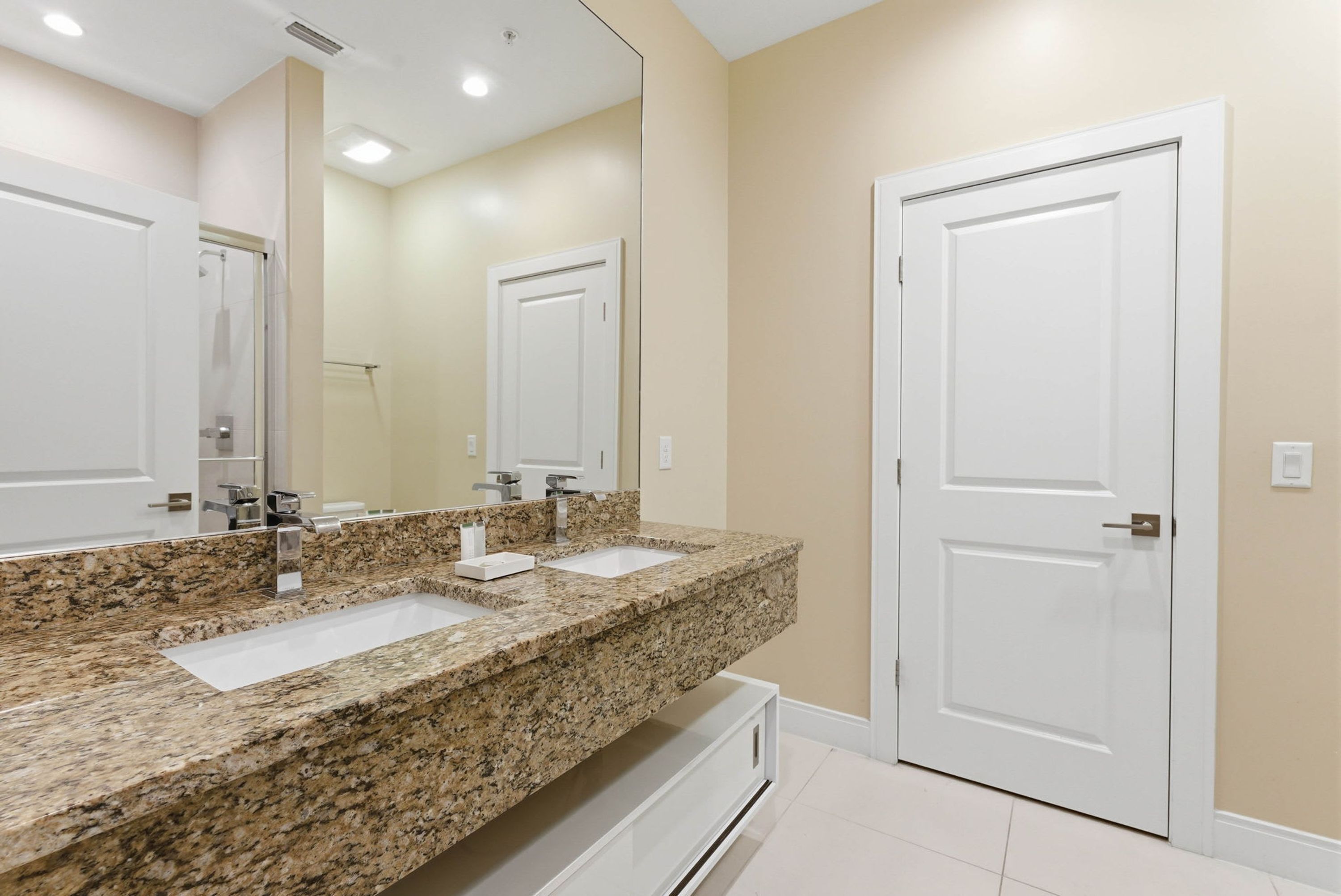 A bathroom with a granite countertop, two sinks, a mirror, and a white door.
