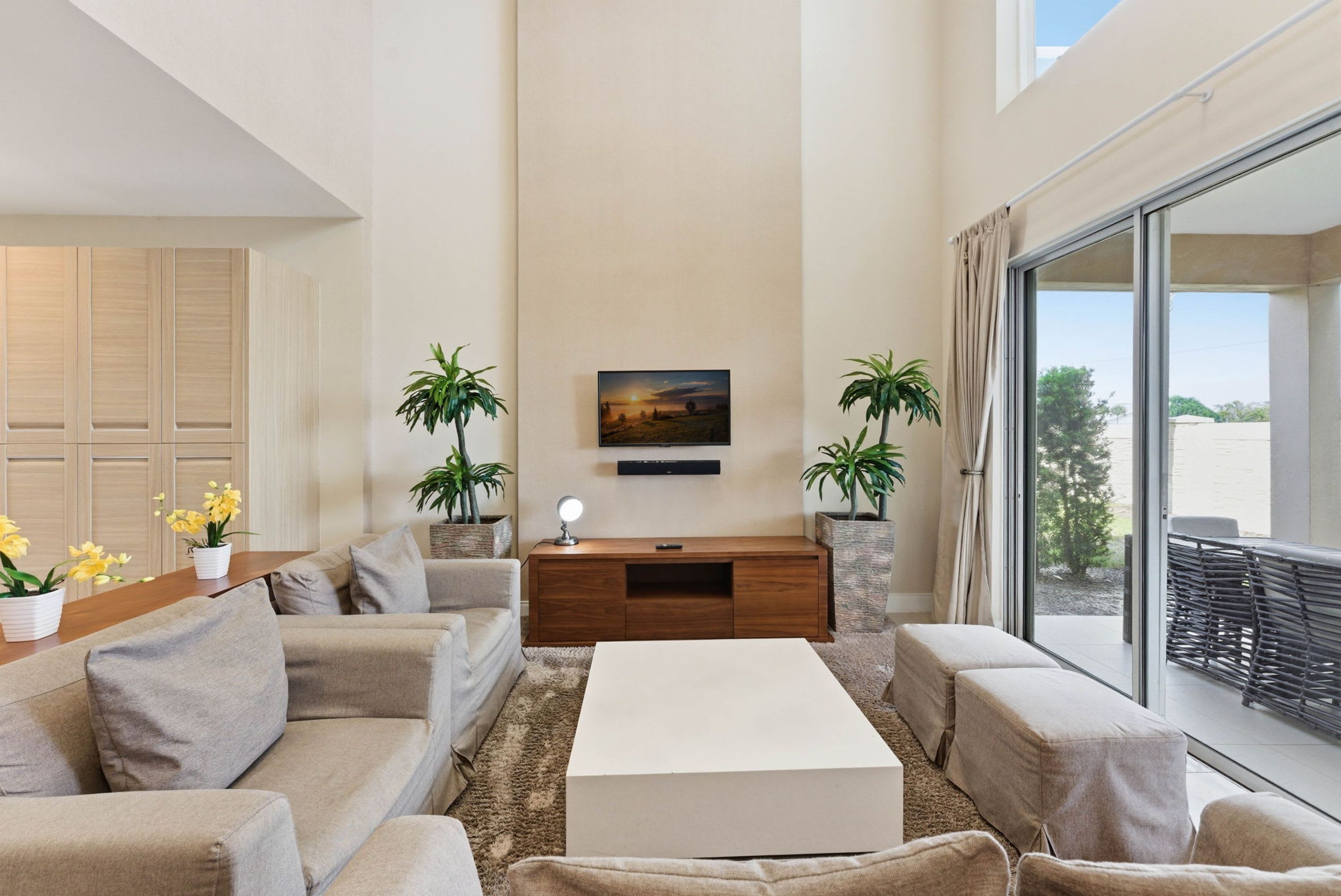 A modern living room with a sofa set, a coffee table, a TV mounted on the wall, and two potted plants.