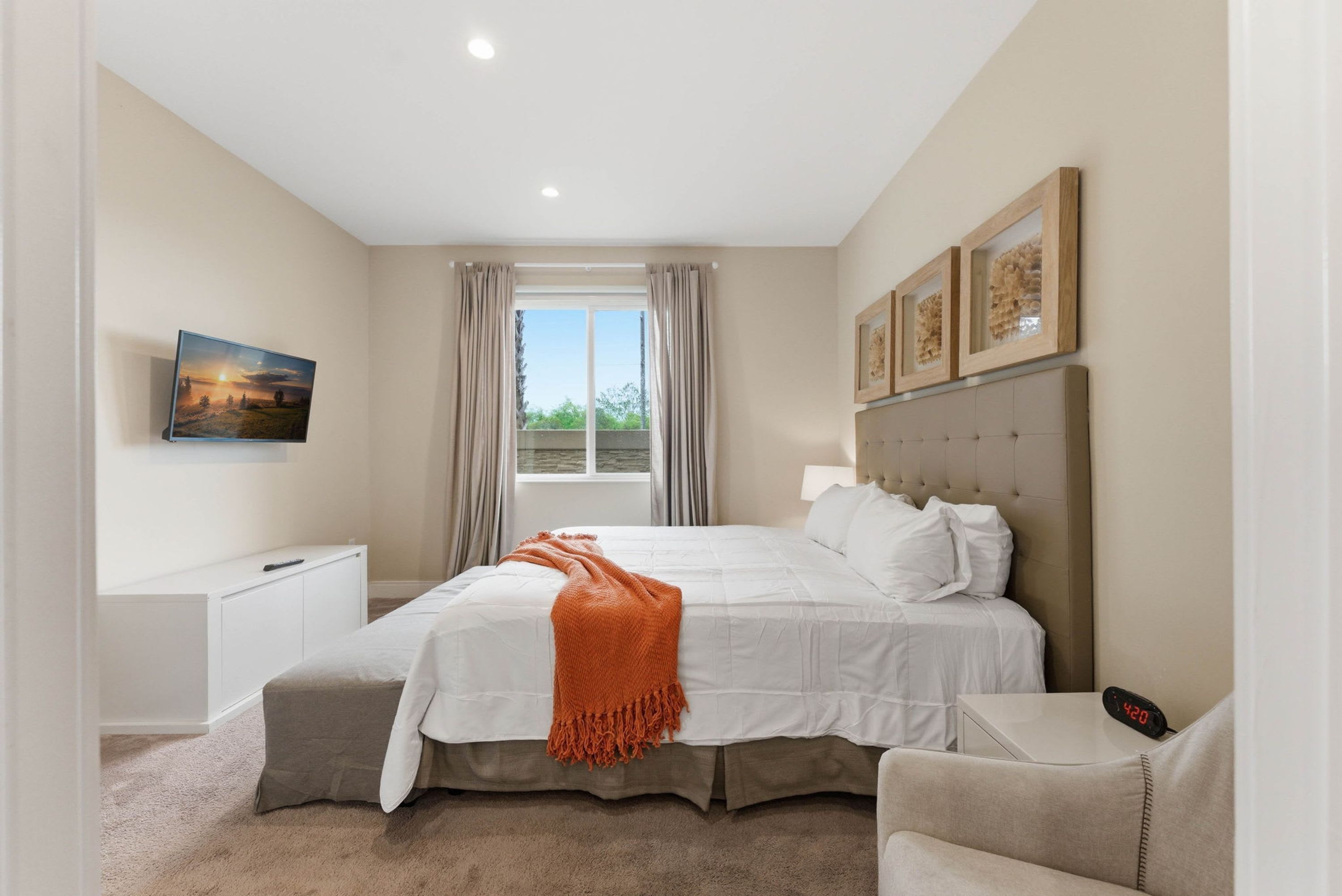 A bedroom with a bed, bedside tables, a chair, a window with curtains, a painting on the wall, and a clock on the nightstand.