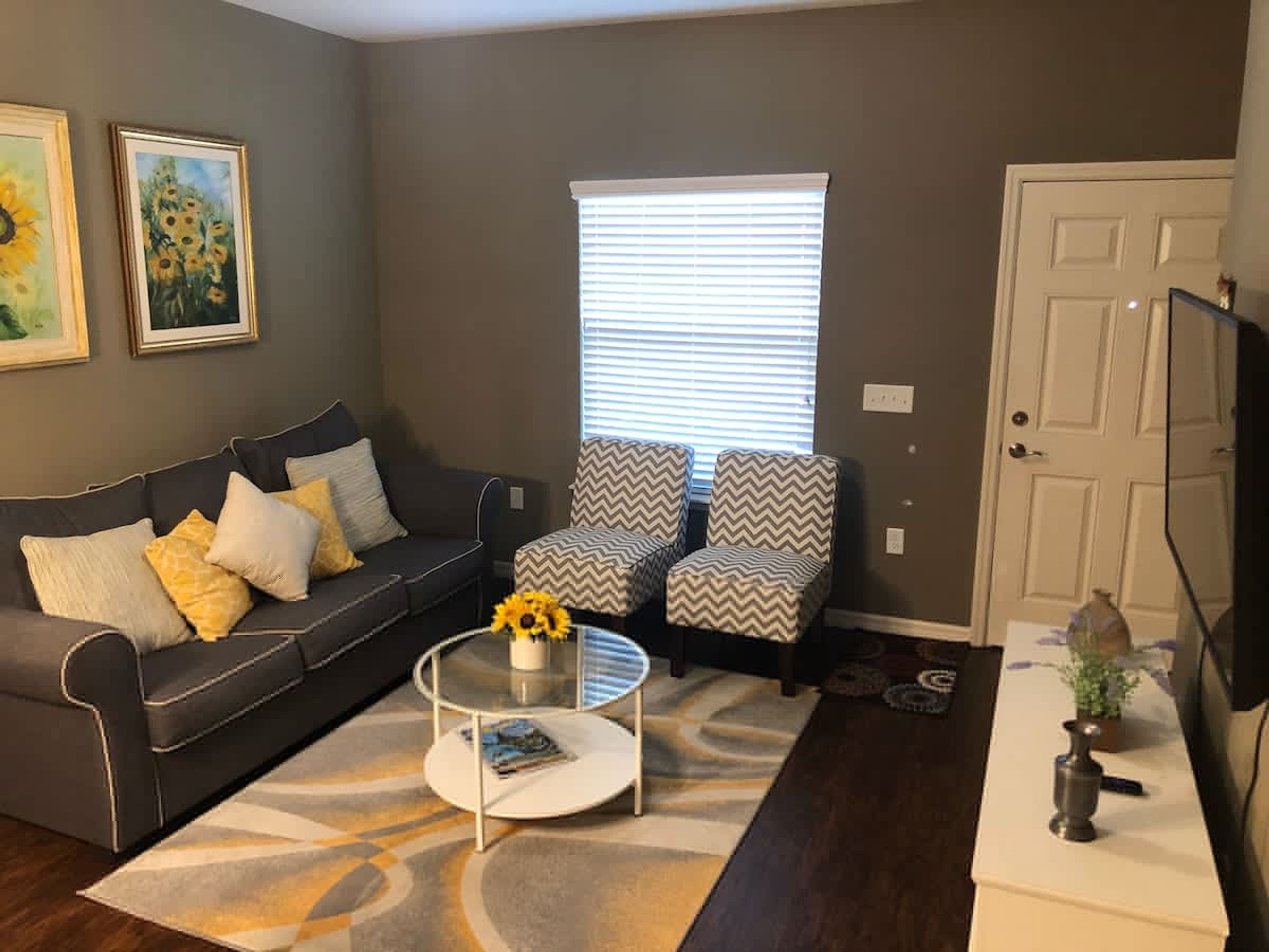 Una sala de estar con un sofá gris, dos sillones estampados, una mesa de centro y un televisor sobre un mueble bajo.