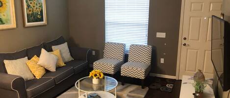 Una sala de estar con un sofá gris, dos sillones estampados, una mesa de centro y un televisor sobre un mueble bajo.