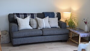 A grey sofa with multiple cushions, a small side table with a lamp, and a framed picture on the wall. - Classic Kiwi Bach + Sleepout - 100m to Purau beach (Purau)