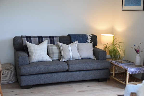 Ein graues Sofa mit mehreren Kissen, ein kleiner Beistelltisch mit Lampe und ein gerahmtes Bild an der Wand.