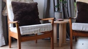 A room with a flat-screen TV mounted on the wall, two wooden chairs with cushions, a small round table with a plant, and grey curtains. - Classic Kiwi Bach + Sleepout - 100m to Purau beach (Purau)