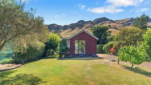A red wooden cabin with a large window, surrounded by a well-maintained lawn and various shrubs, set against a backdrop of mountains. - Classic Kiwi Bach + Sleepout - 100m to Purau beach (Purau)