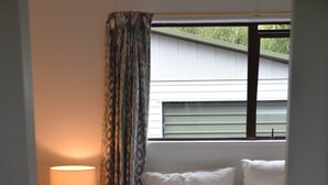 A bedroom with a bed, pillows, a bedside lamp, a nightstand, and a window with curtains. - Classic Kiwi Bach + Sleepout - 100m to Purau beach (Purau)