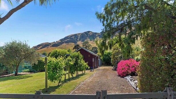 A gravel path leading to a red building, surrounded by greenery and a mountain in the background. - Classic Kiwi Bach + Sleepout - 100m to Purau beach (Purau)