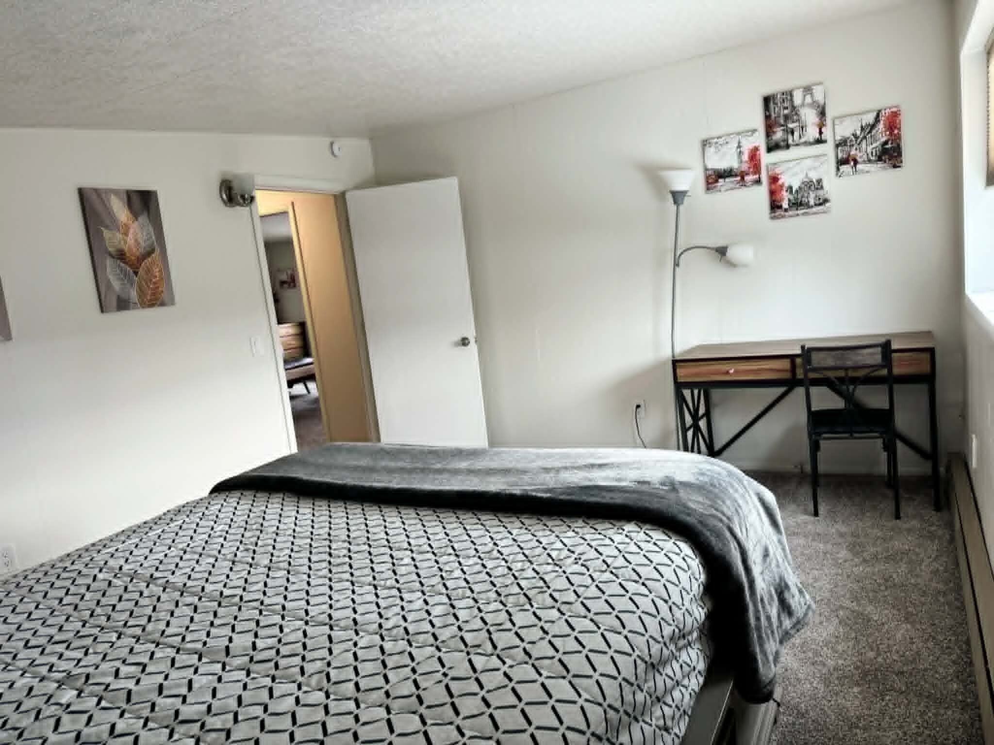 A bedroom with a bed, a desk, and a chair. There are wall art pieces and a door leading to another room.
