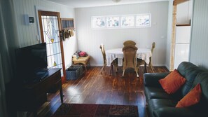 A living room with a television, a black leather sofa, and a dining area with wicker chairs and a table. - The Gardener's Cottage (Warrentinna)