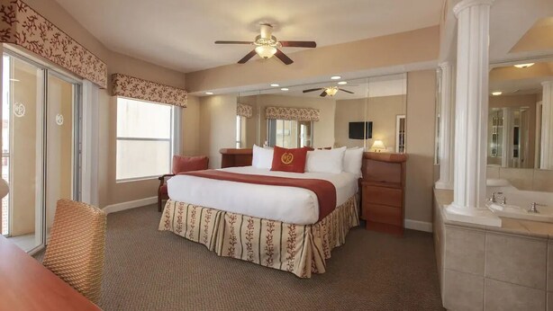 A hotel room with a large bed, a desk, a chair, a ceiling fan, a mirror, and a bathroom with a sink and mirror. - Westgate town center resort / 2 bedrooms (Kissimmee)