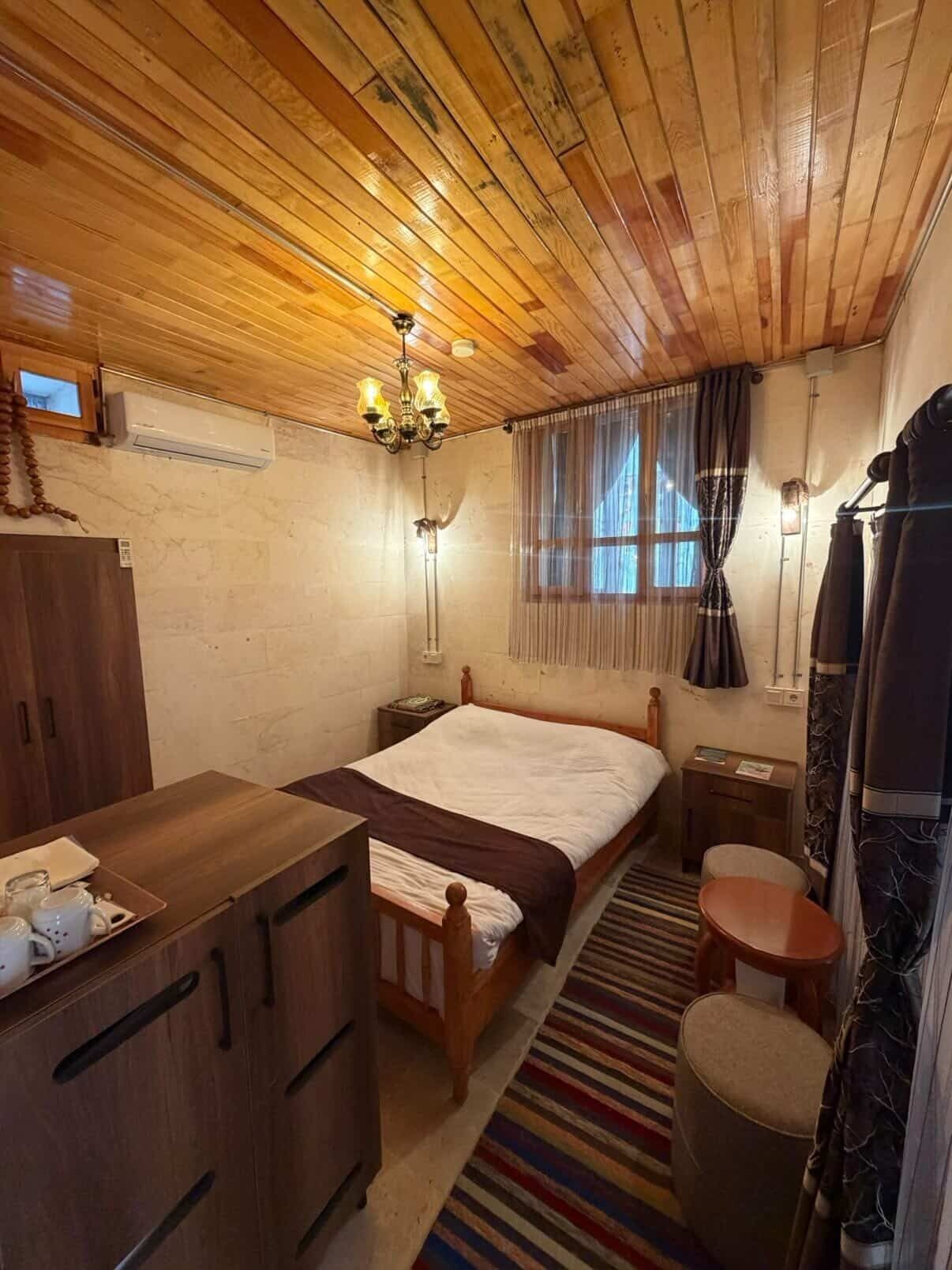 A wooden-paneled room with a bed, bedside table, chair, and a small desk with drawers.