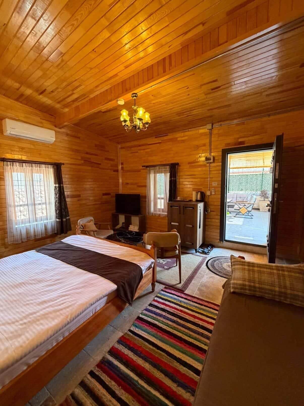 Habitación de cabaña de madera con una cama, un escritorio, una silla, un televisor y un ventanal corredera hacia el exterior.