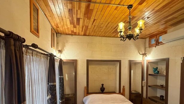 A bedroom with a wooden ceiling, a bed, a chair, a small table, and a nightstand. - Ela Sultan Han (Gaziantep)