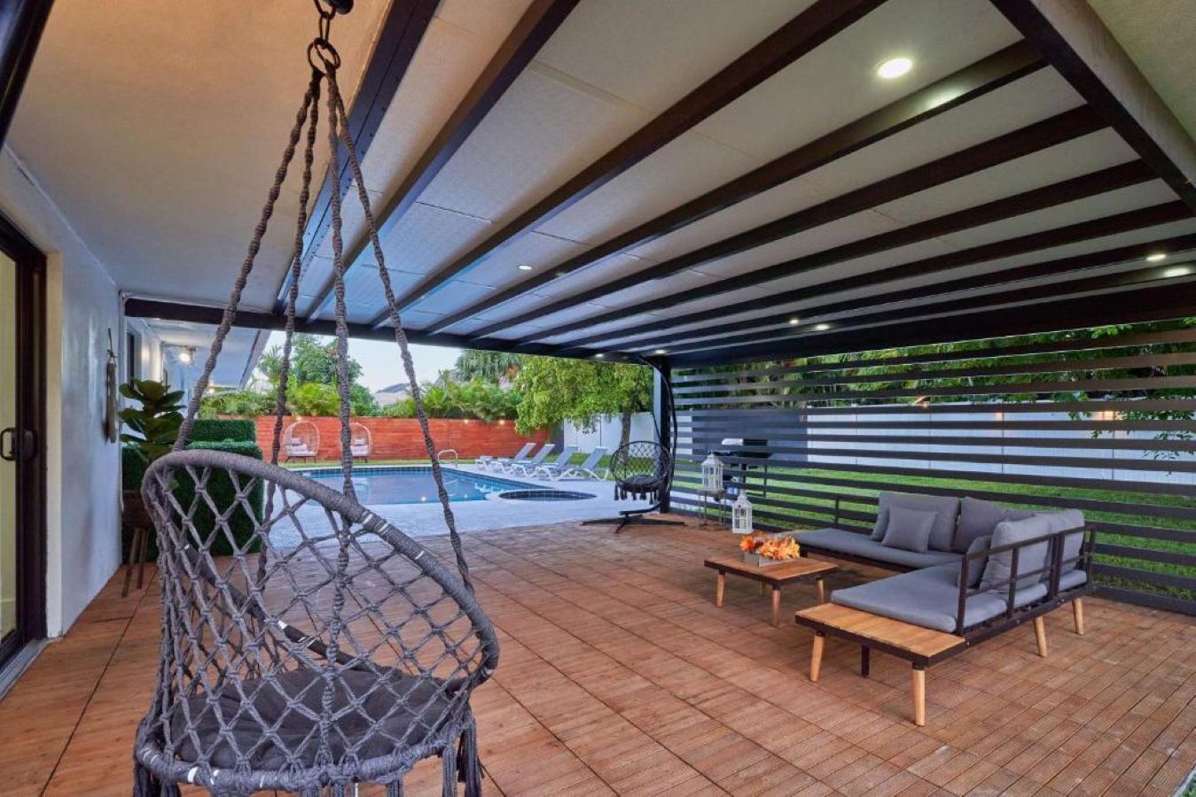 Un espace extérieur abrité comprenant un fauteuil suspendu, un canapé, une petite table et une piscine en arrière-plan.