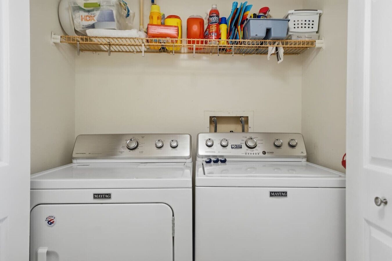 A laundry area with two Maytag washers and dryers, a shelf with cleaning supplies, and a small cabinet.