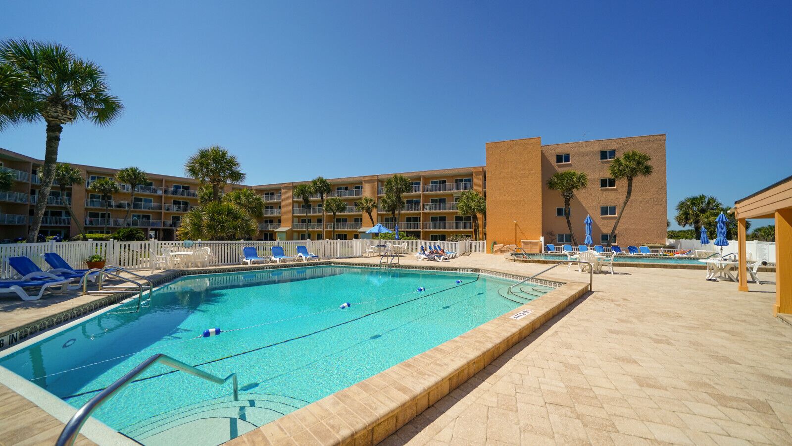 Une piscine avec des chaises longues, une clôture blanche et un immeuble à plusieurs étages en arrière-plan.